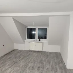 Poddasze po remoncie z szarymi panelami podłogowymi imitującymi drewno, białe ściany i grzejnik pod oknem.