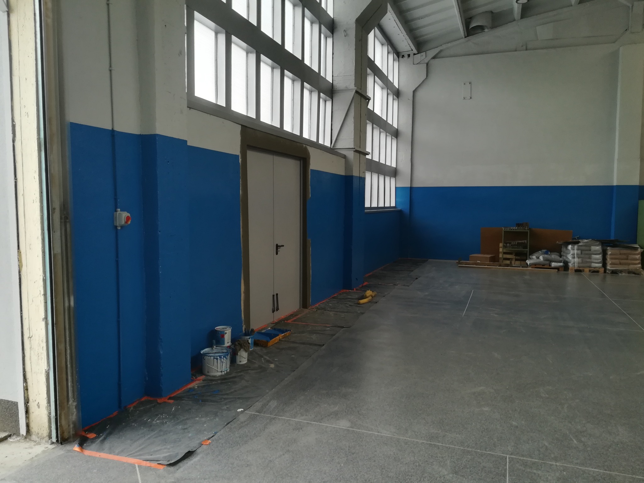 Wnętrze przemysłowe z pomalowaną na niebiesko dolną częścią ścian, widoczne puszki z farbą, zabezpieczona folią podłoga przy ścianie, okna w górnej części ściany, palety i worki w tle.