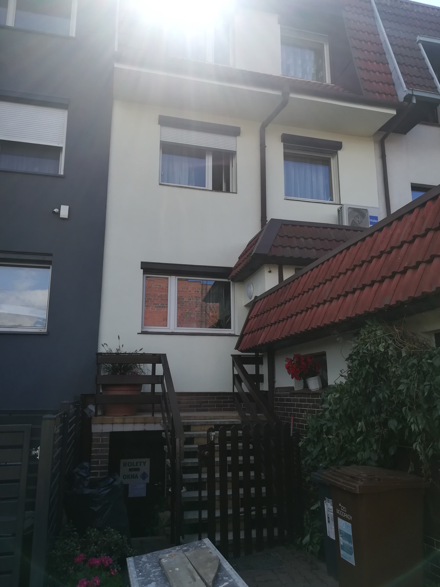 Dwukondygnacyjny dom z jasną elewacją i roletami zewnętrznymi, widoczny z zewnątrz, z fragmentem ciemnoszarej ściany sąsiedniego budynku po lewej stronie, czerwonym dachem i zielenią w ogrodzie.