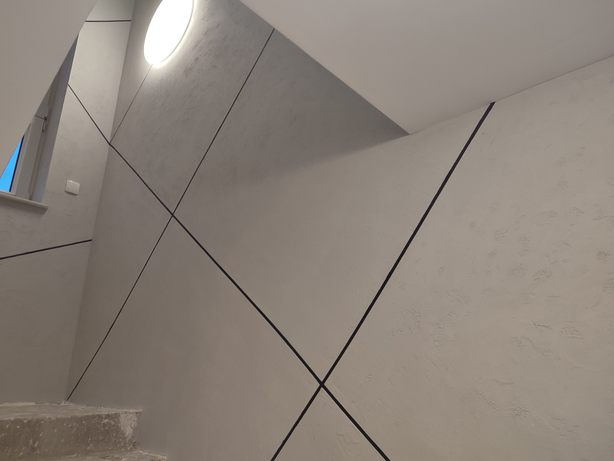 Klatka schodowa z wykończonymi ścianami w odcieniach szarości, z dekoracyjnymi czarnymi listwami tworzącymi geometryczne wzory i widocznym fragmentem okna.