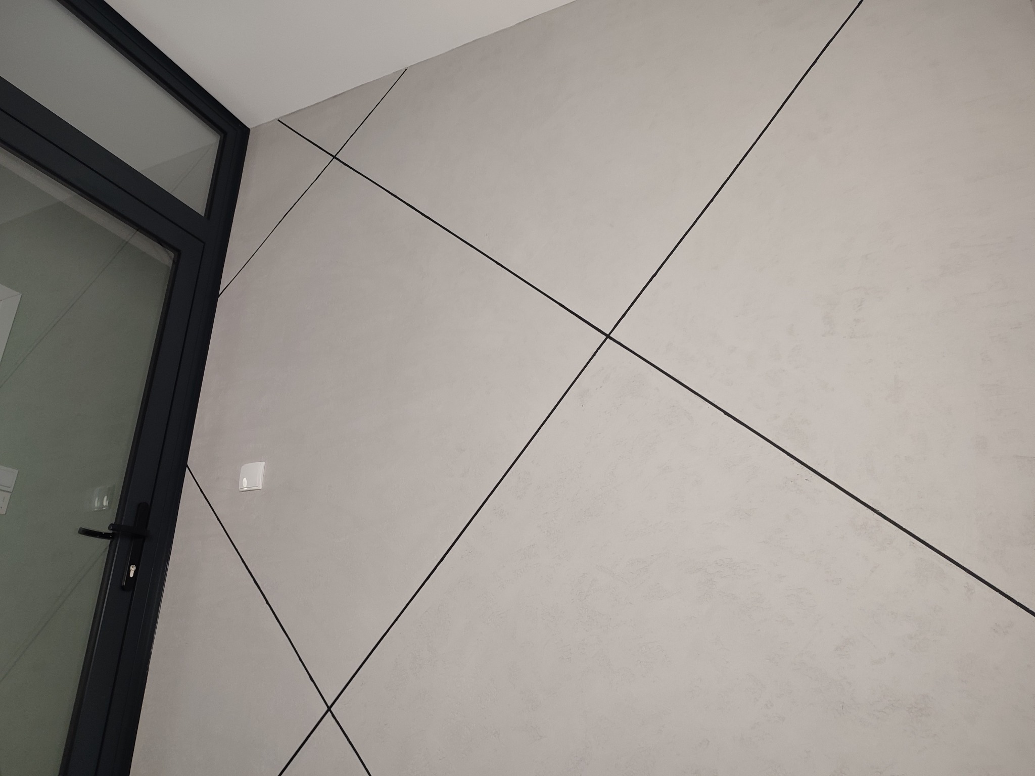 Ściana z dekoracyjnym tynkiem w odcieniach szarości, z czarnymi liniami tworzącymi geometryczny wzór, widoczny fragment czarnej ramy drzwi i biały włącznik światła.
