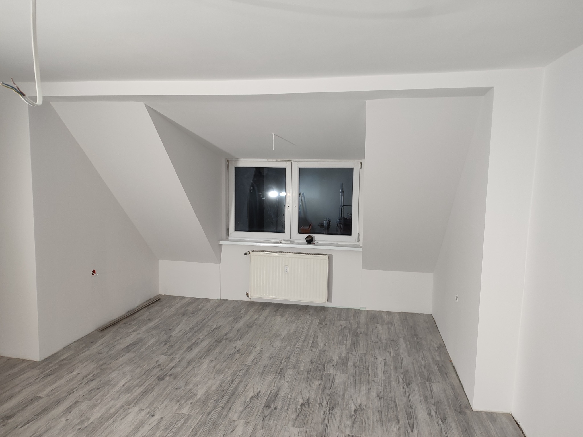 Poddasze po remoncie z szarymi panelami podłogowymi imitującymi drewno, białe ściany i grzejnik pod oknem.
