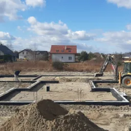 Przygotowanie terenu pod fundamenty domu: wykopane rowy, zalane betonem, widoczna izolacja, wystające pręty zbrojeniowe, w tle budynki mieszkalne i pracujący operator koparko-ładowarki Volvo.