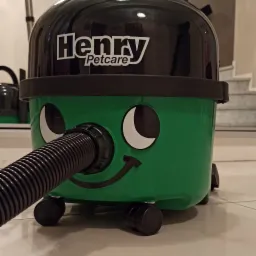 Zielony odkurzacz Henry Petcare z charakterystycznym uśmiechem i wężem ssącym, stojący na błyszczącej podłodze, z widocznymi schodami w tle.