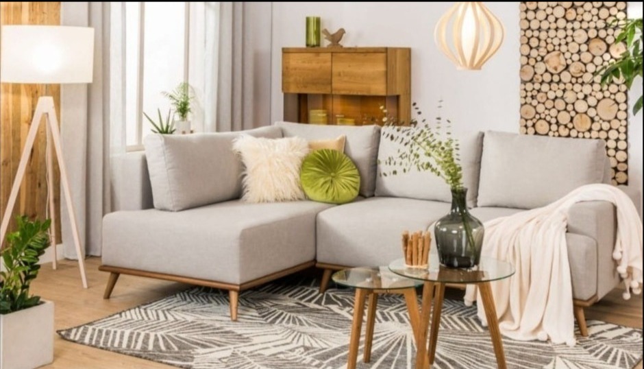 Jasnoszara narożna kanapa z poduszkami i pledem w salonie z drewnianymi akcentami, lampą trójnożną i stolikiem kawowym na wzorzystym dywanie.