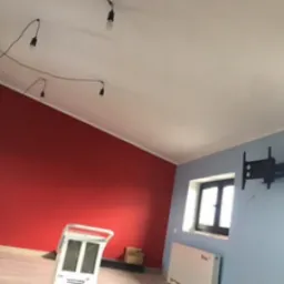 Pokój po malowaniu, z czerwoną ścianą, jasnoniebieską ścianą z zamontowanym uchwytem na telewizor, widoczne białe okno, grzejnik i lampa wisząca z wieloma kablami.