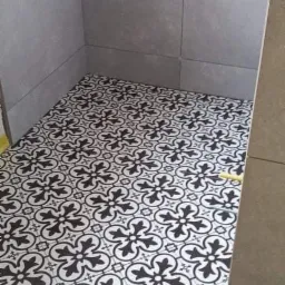 Ułożona podłoga z płytek ceramicznych w czarno-biały wzór kwiatowy w rogu łazienki z szarymi płytkami ściennymi.