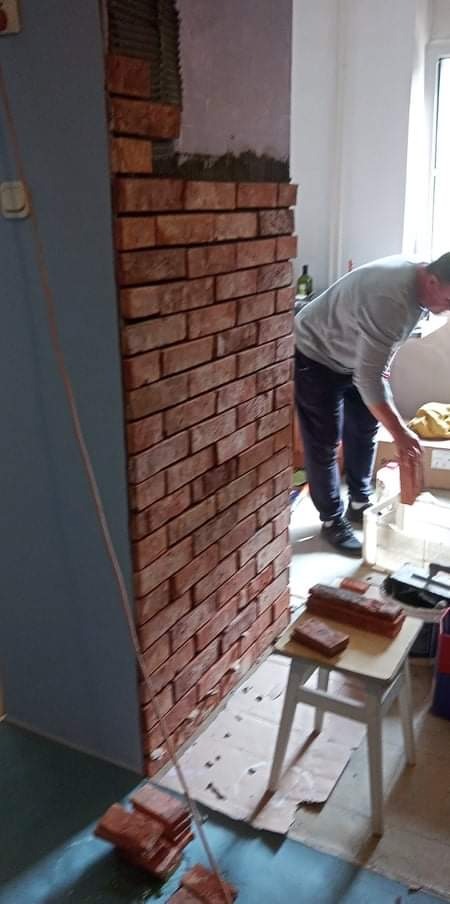 Montaż cegieł dekoracyjnych na ścianie w pomieszczeniu, widoczny fragment ściany z ułożonymi cegłami, pracownik w trakcie układania, biały taboret z cegłami obok.