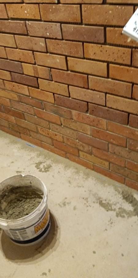 Ściana wyłożona cegłą klinkierową w trakcie prac, widoczne wiadro z zaprawą murarską i narzędzie do spoinowania w prawym górnym rogu.