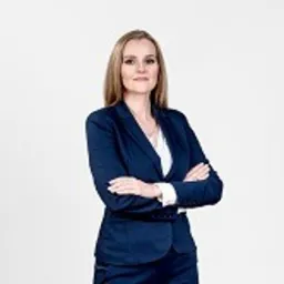 Portret kobiety w granatowym garniturze, stojącej na jasnym tle z założonymi rękami.