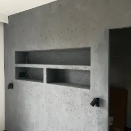 Ściana z szarą, dekoracyjną fakturą tynku, wnęki z półkami, gniazdka elektryczne i dwa czarne reflektory punktowe.