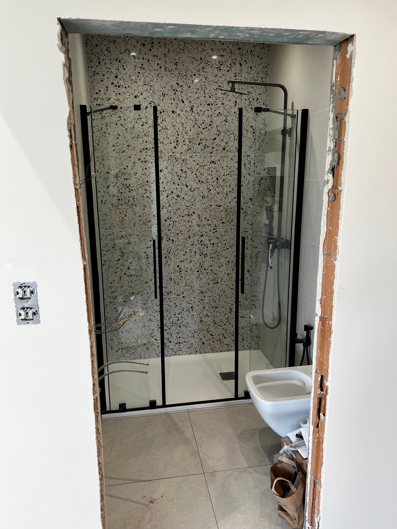 Nowoczesna kabina prysznicowa z czarnymi ramami i deszczownicą, widoczna przez surowy otwór drzwiowy w trakcie remontu łazienki, z widocznymi przewodami elektrycznymi i częściowo wykończonymi...