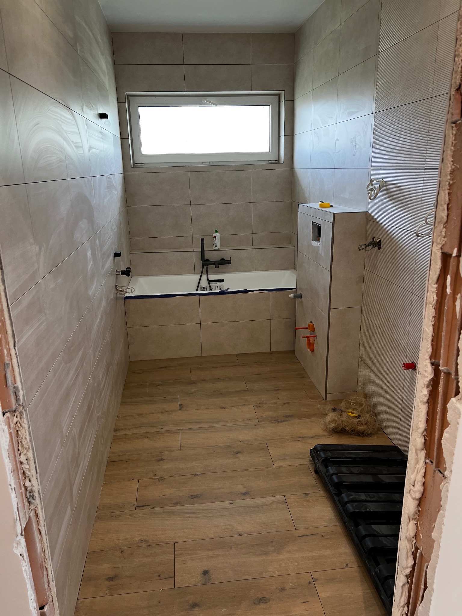 Wnętrze łazienki w trakcie remontu: ułożone płytki ścienne w odcieniach beżu, drewnopodobne płytki na podłodze, zabudowana wanna, fragment ściany przed wykończeniem, widoczne instalacje...