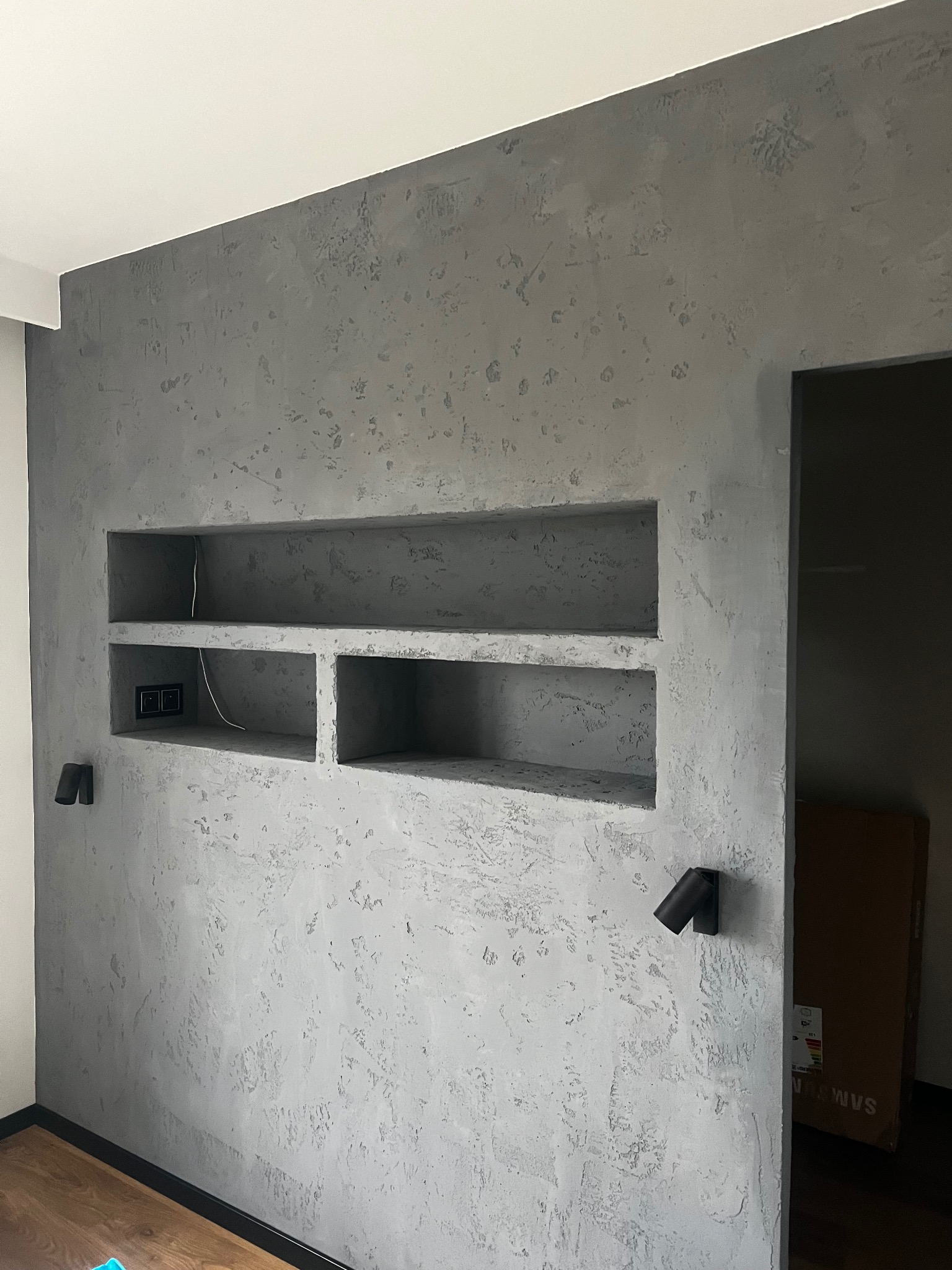 Ściana z szarą, dekoracyjną fakturą tynku, wnęki z półkami, gniazdka elektryczne i dwa czarne reflektory punktowe.