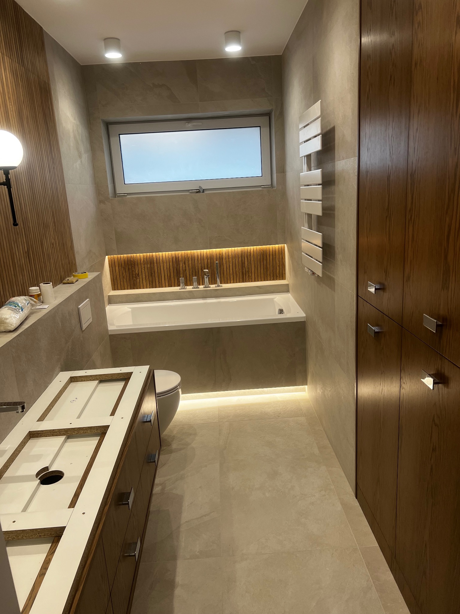 Wnętrze łazienki w trakcie prac wykończeniowych, widoczna wanna z podświetleniem LED, grzejnik drabinkowy, zabudowa szafkowa z drewnopodobnymi frontami oraz częściowo zmontowana szafka pod umywalkę.