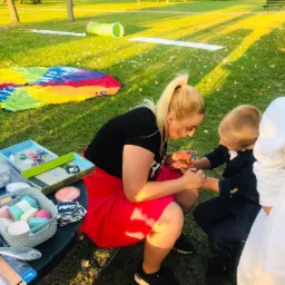 Kobieta w czerwonej spódnicy maluje brokatowy wzór na ręce chłopca w granatowym garniturze, na trawie rozłożone kolorowe rekwizyty i tunel dla dzieci.