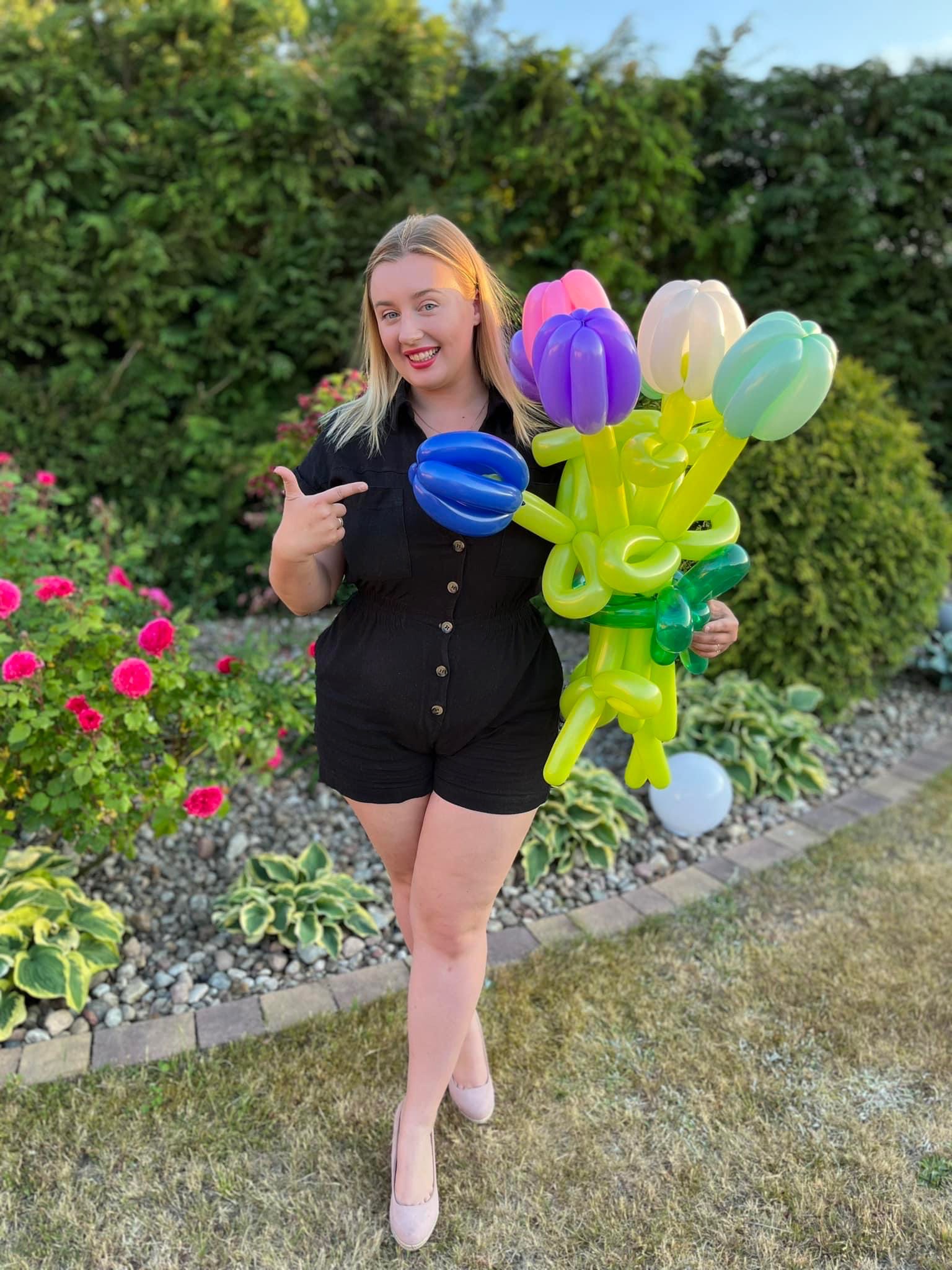 Uśmiechnięta kobieta w czarnym kombinezonie prezentuje bukiet z balonów w kształcie tulipanów (różowy, fioletowy, biały, zielony) na tle zielonego żywopłotu i kwitnących róż.