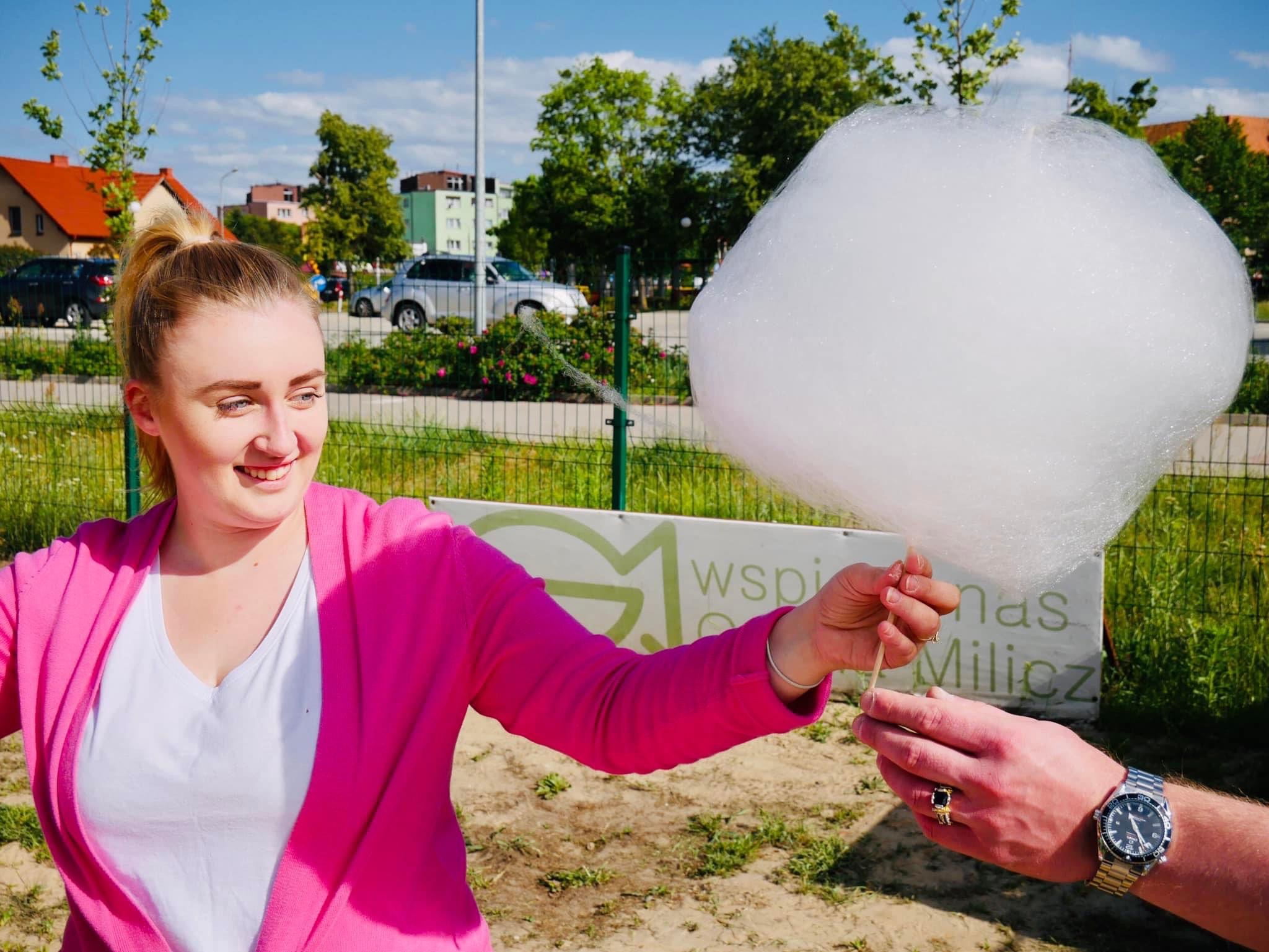 Uśmiechnięta blondynka w różowym swetrze podaje watę cukrową, trzymaną przez męską dłoń z zegarkiem Omega Seamaster i sygnetem na tle zieleni i budynków.
