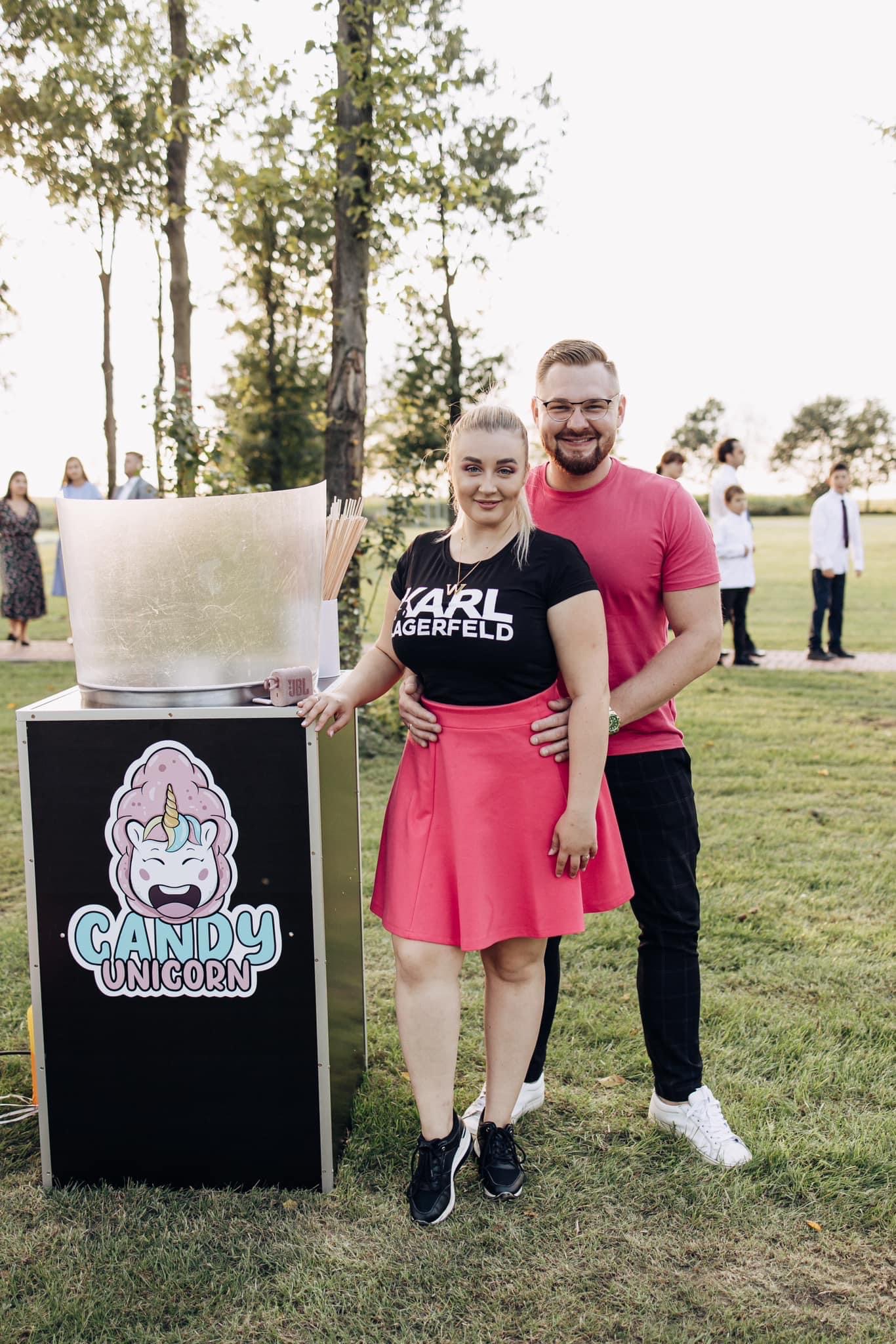 Para prezentuje wózek do waty cukrowej z logo 'Candy Unicorn' na tle zielonej trawy i drzew, w tle widoczni goście.