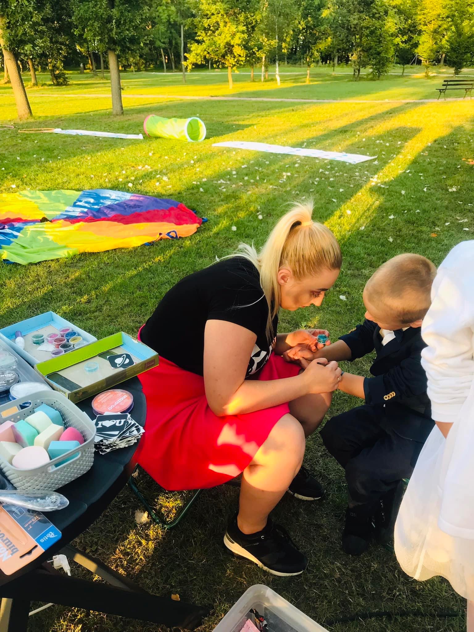 Kobieta w czerwonej spódnicy maluje brokatowy wzór na ręce chłopca w granatowym garniturze, na trawie rozłożone kolorowe rekwizyty i tunel dla dzieci.