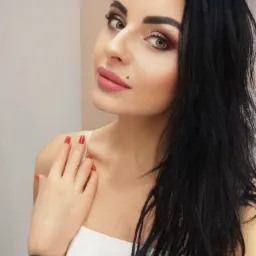 Portret kobiety z ciemnymi włosami, delikatnym makijażem, czerwonymi paznokciami i tatuażem na ramieniu, pozującej na jasnym tle.