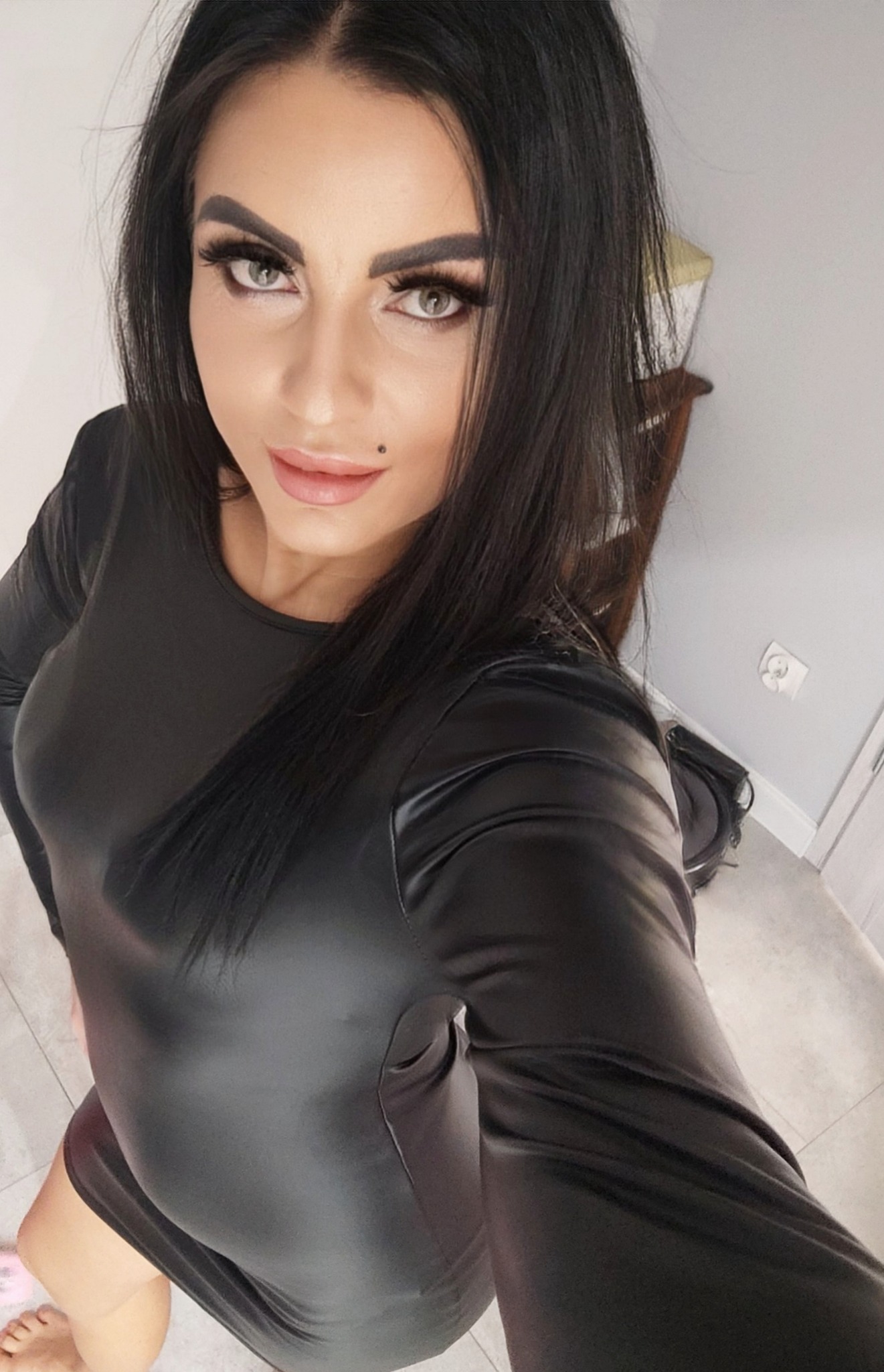 Kobieta z ciemnymi włosami i mocnym makijażem w czarnej, obcisłej sukience pozuje do selfie, z widocznym znamieniem na ustach.