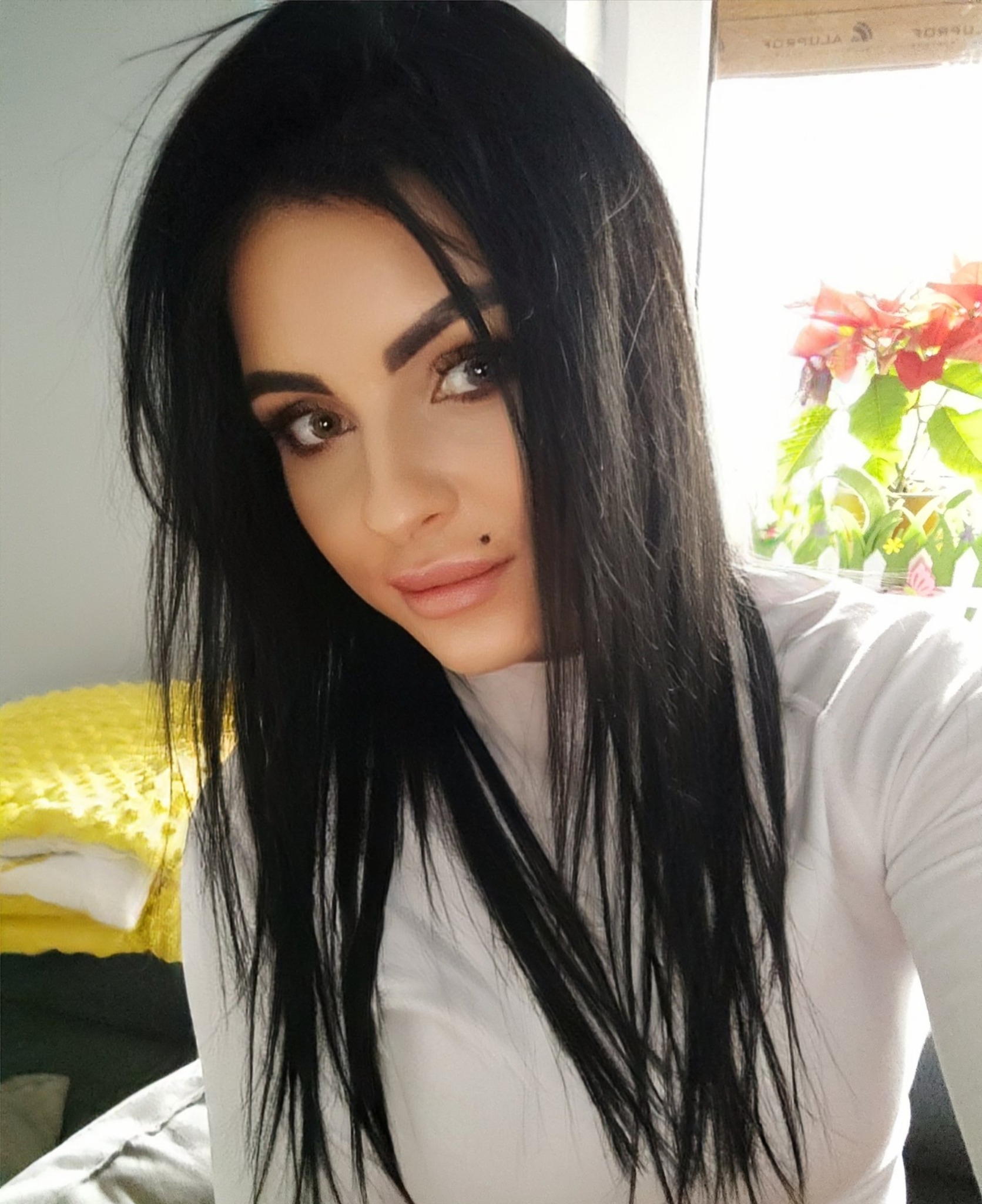 Portret kobiety z długimi, czarnymi włosami, z widocznym znamieniem na twarzy, delikatny makijaż oczu, w tle jasne okno z rośliną.