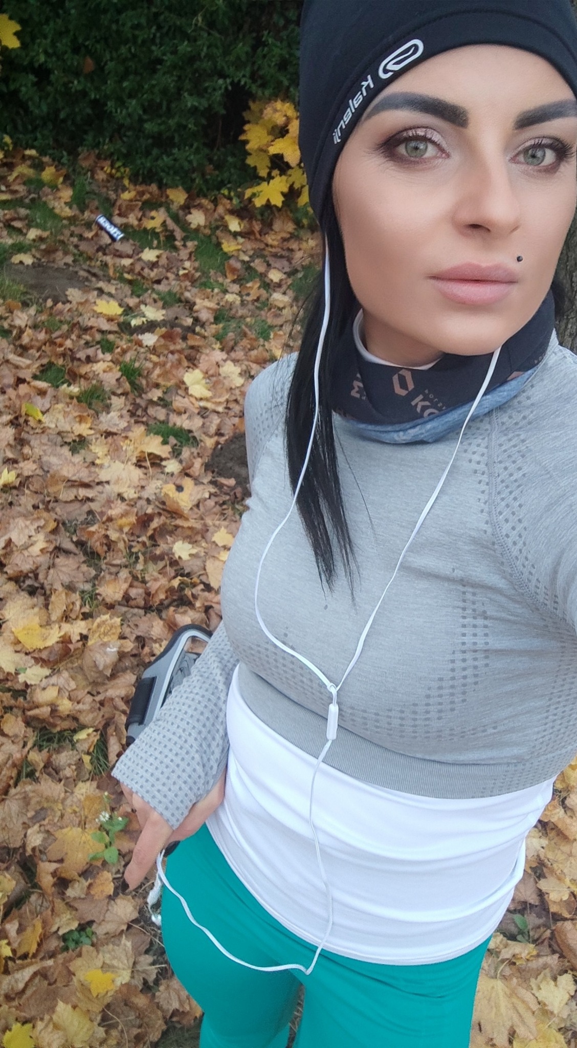 Kobieta w sportowym stroju, czapce i słuchawkach, robiąca selfie na tle jesiennych liści w parku.