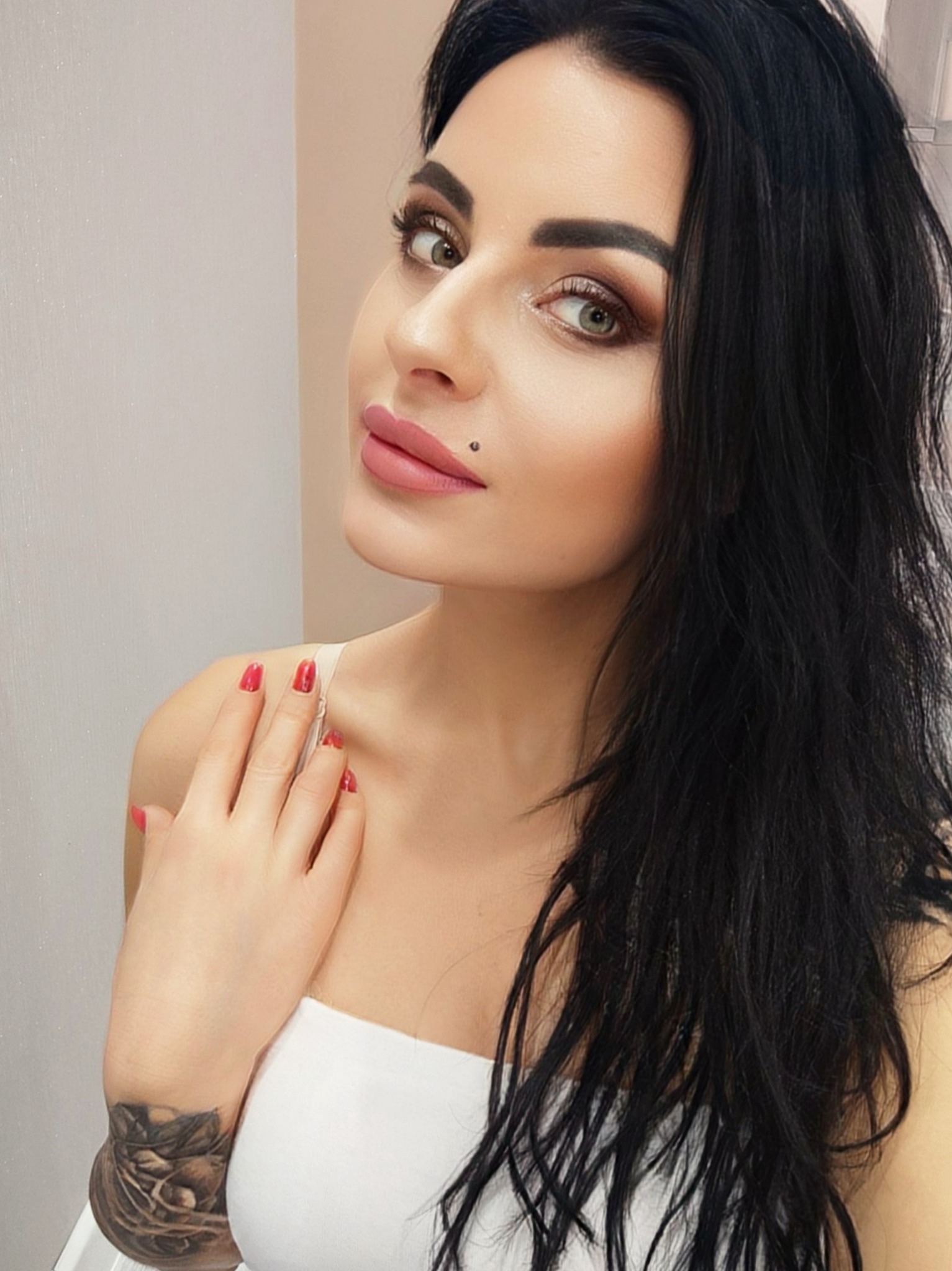 Portret kobiety z ciemnymi włosami, delikatnym makijażem, czerwonymi paznokciami i tatuażem na ramieniu, pozującej na jasnym tle.