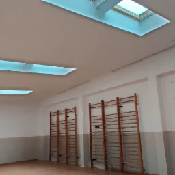 Wnętrze sali gimnastycznej po odświeżeniu, z widocznymi drabinkami gimnastycznymi, pomalowanymi ścianami w dwóch odcieniach szarości i bieli oraz trzema świetlikami w suficie, z których jeden to...