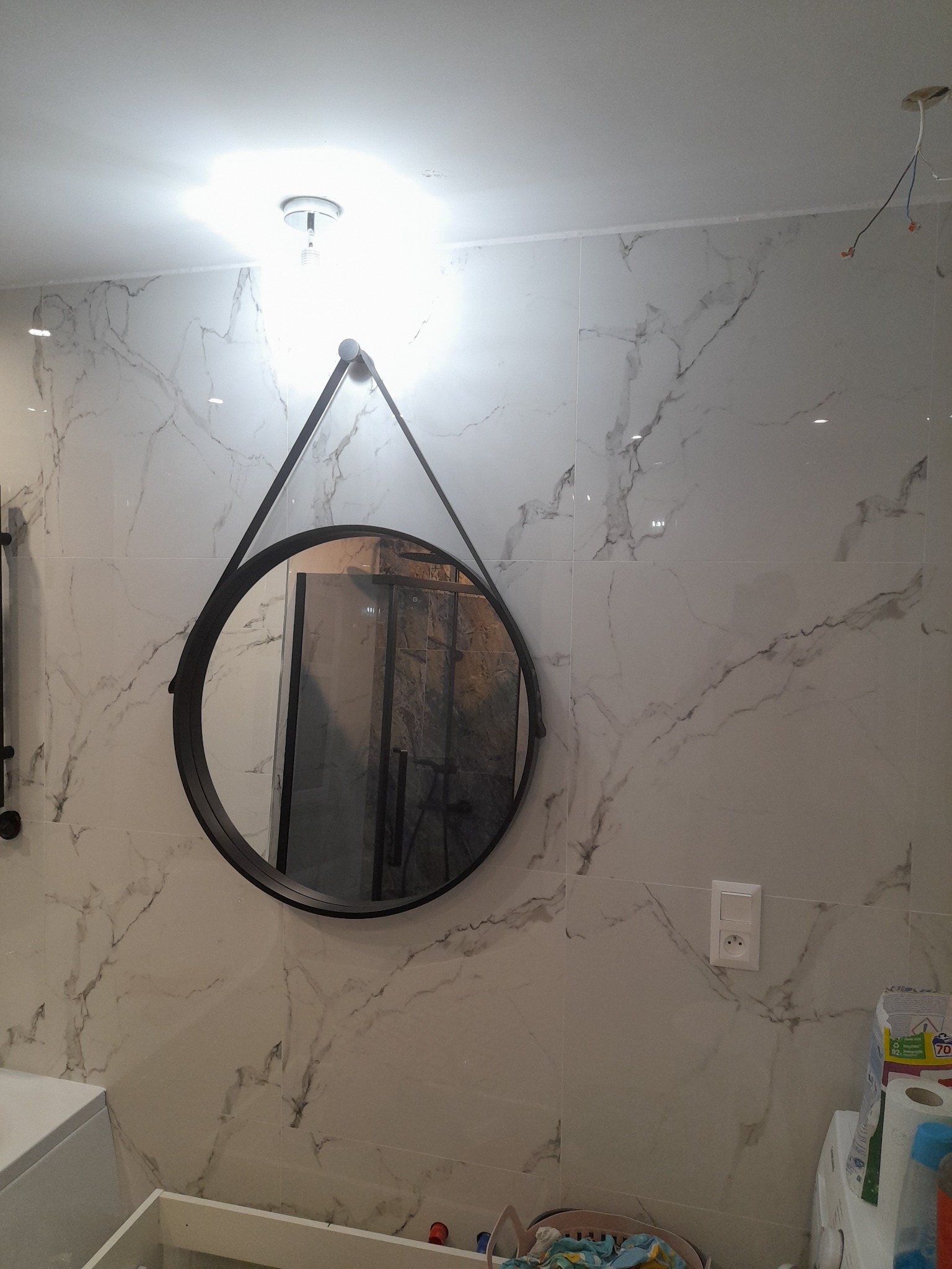 Okrągłe lustro w czarnej ramie zawieszone na ścianie wyłożonej białymi płytkami imitującymi marmur. Widoczne gniazdko elektryczne, fragment wanny i instalacja elektryczna w suficie.