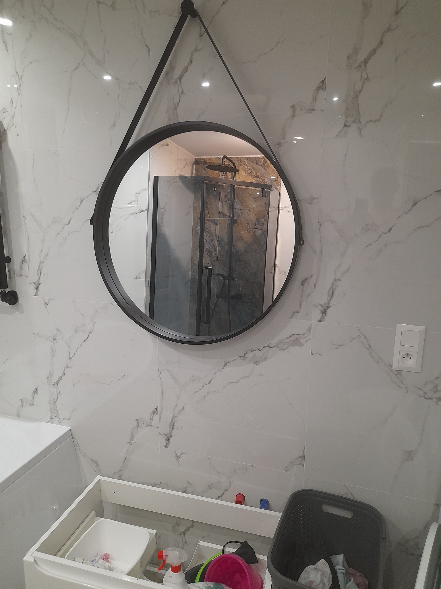 Okrągłe lustro w czarnej ramie zawieszone na marmurowej ścianie w łazience, odbijające kabinę prysznicową. Pod lustrem biała szafka pod umywalkę z widocznymi środkami czystości i koszem na pranie.