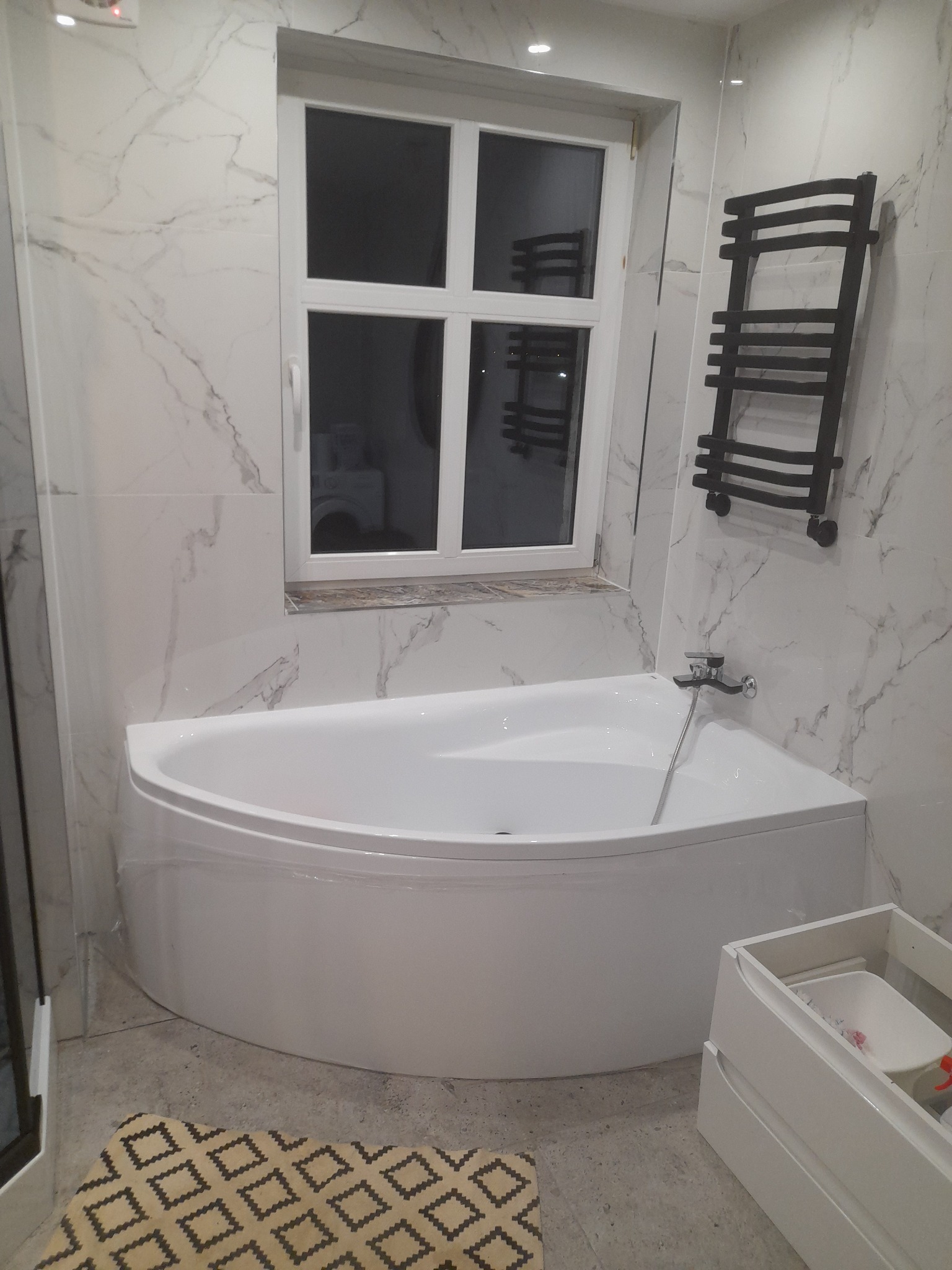 Biała wanna narożna pod oknem w wykończonej łazience w Sławnie, marmurowe płytki na ścianach, grzejnik drabinkowy i szafka pod umywalkę.