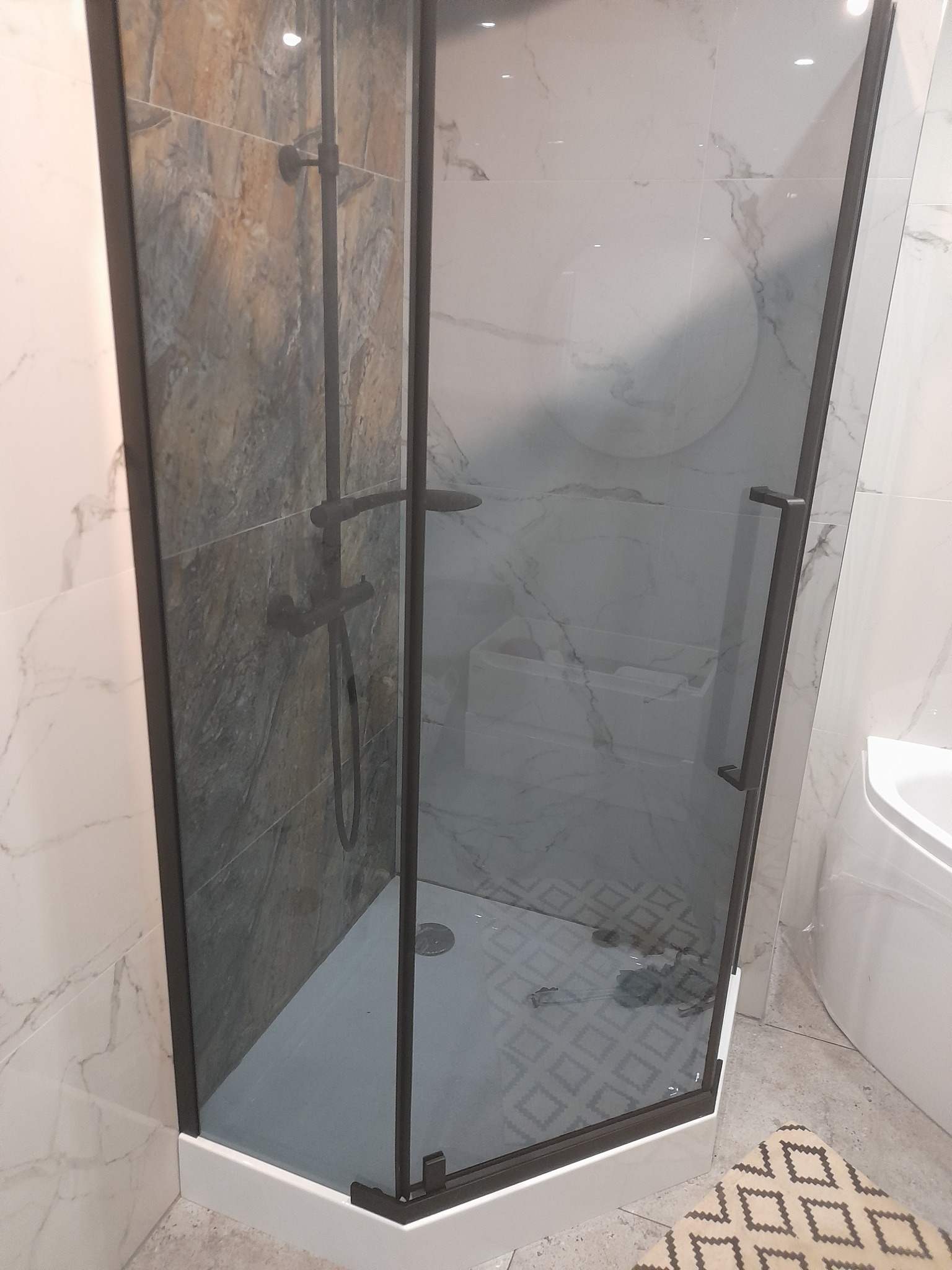 Kabina prysznicowa z czarną armaturą i szklanymi drzwiami, widoczna fragment ściany z płytek imitujących kamień i podłoga z geometrycznym wzorem, w tle wanna.