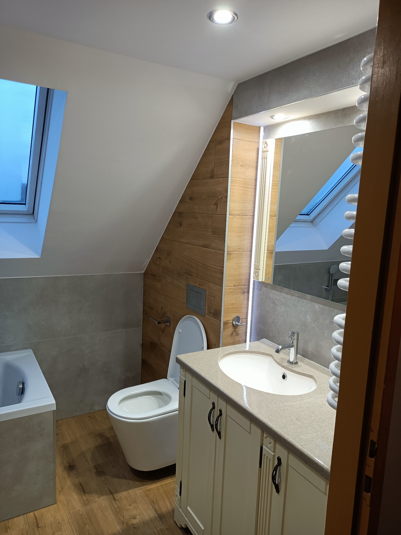 Wykończona łazienka na poddaszu w Berlinie z oknem dachowym, drewnopodobnymi płytkami na ścianie i podłodze, oraz białymi meblami łazienkowymi z umywalką i toaletą typu Geberit.