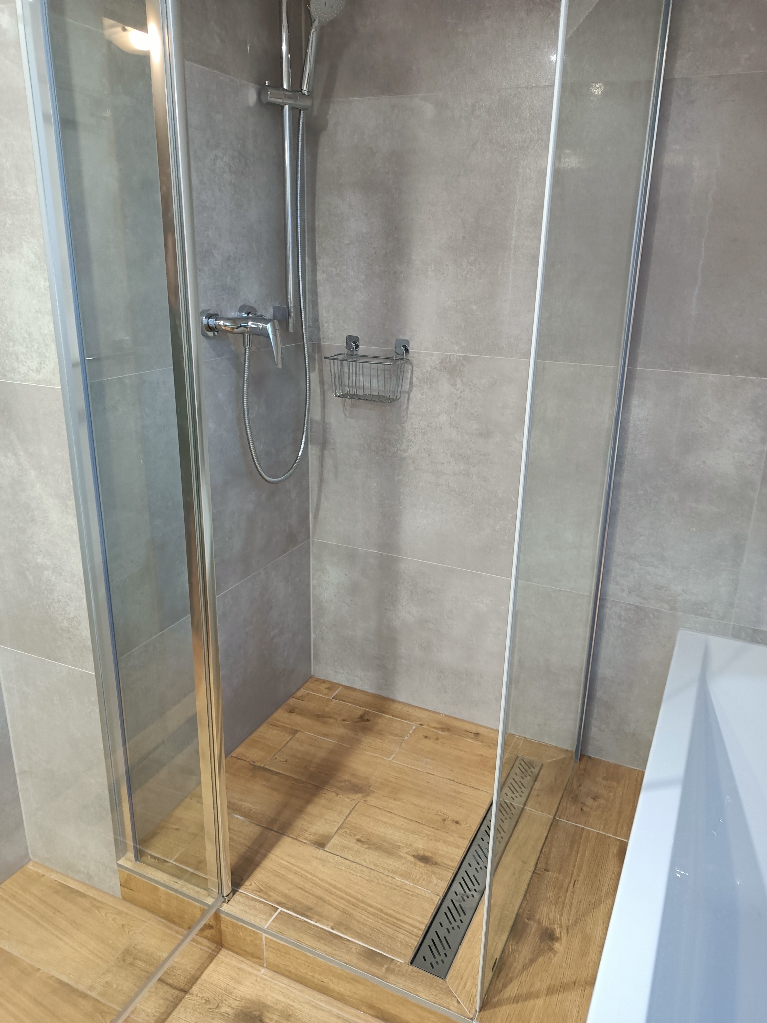 Nowoczesna kabina prysznicowa z drewnopodobną podłogą i szarymi płytkami ściennymi, widoczny odpływ liniowy i chromowana armatura.