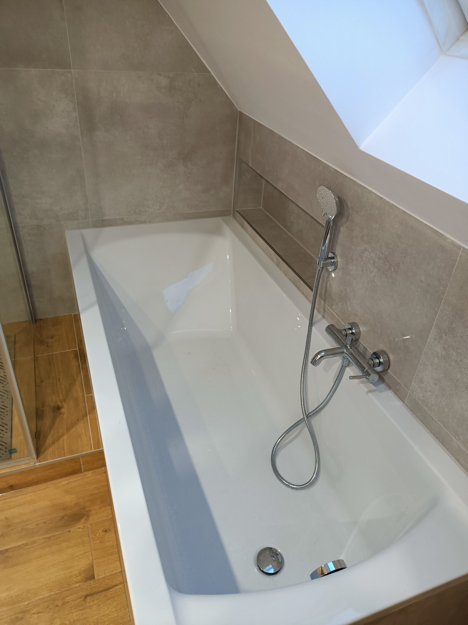 Wanna w łazience na poddaszu, obłożona szarymi płytkami, z armaturą chromowaną i drewnopodobną podłogą, oświetlona światłem wpadającym przez okno dachowe.