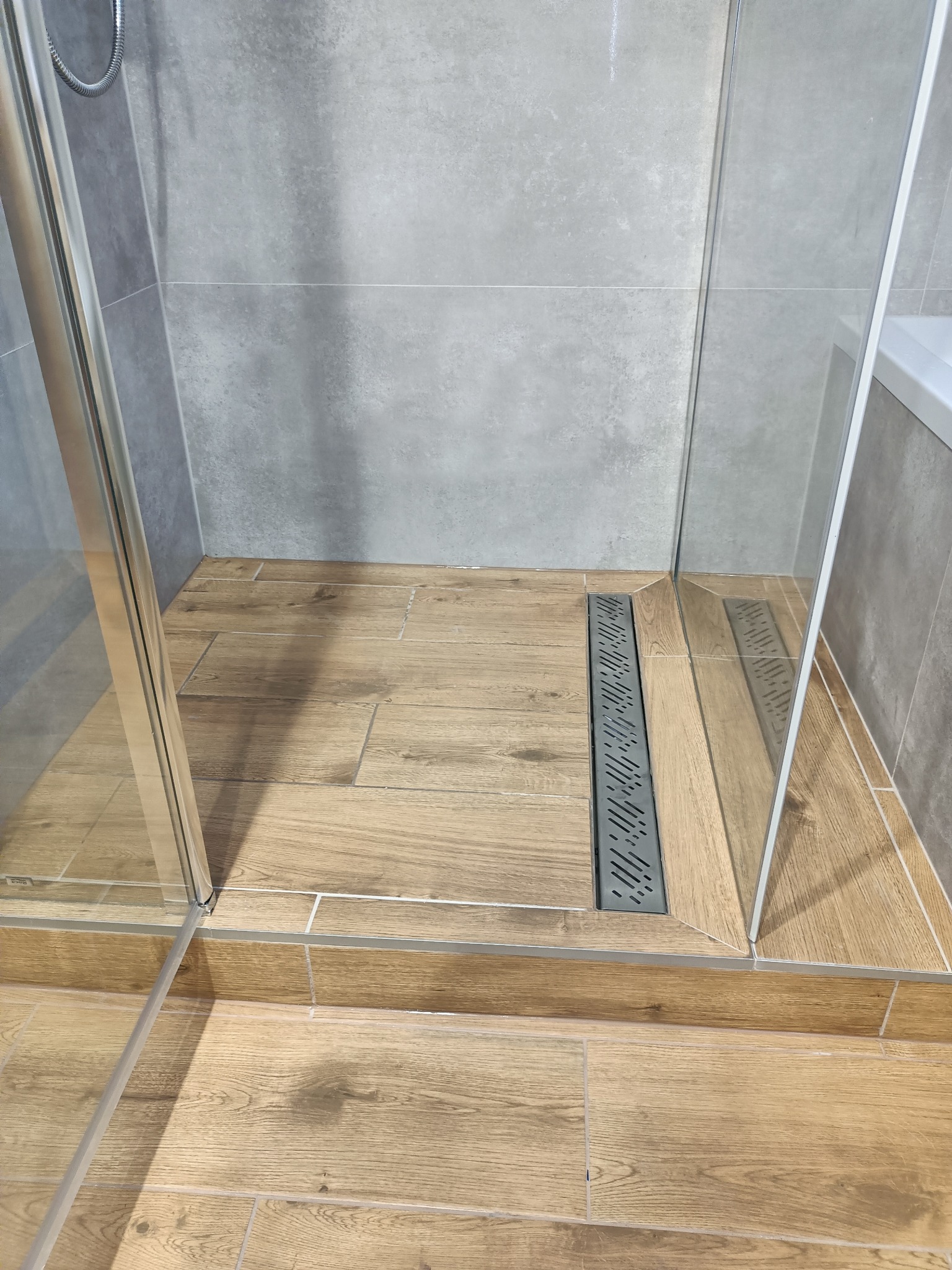 Kabina prysznicowa z odpływem liniowym w podłodze wyłożonej płytkami imitującymi drewno, szare płytki na ścianach.