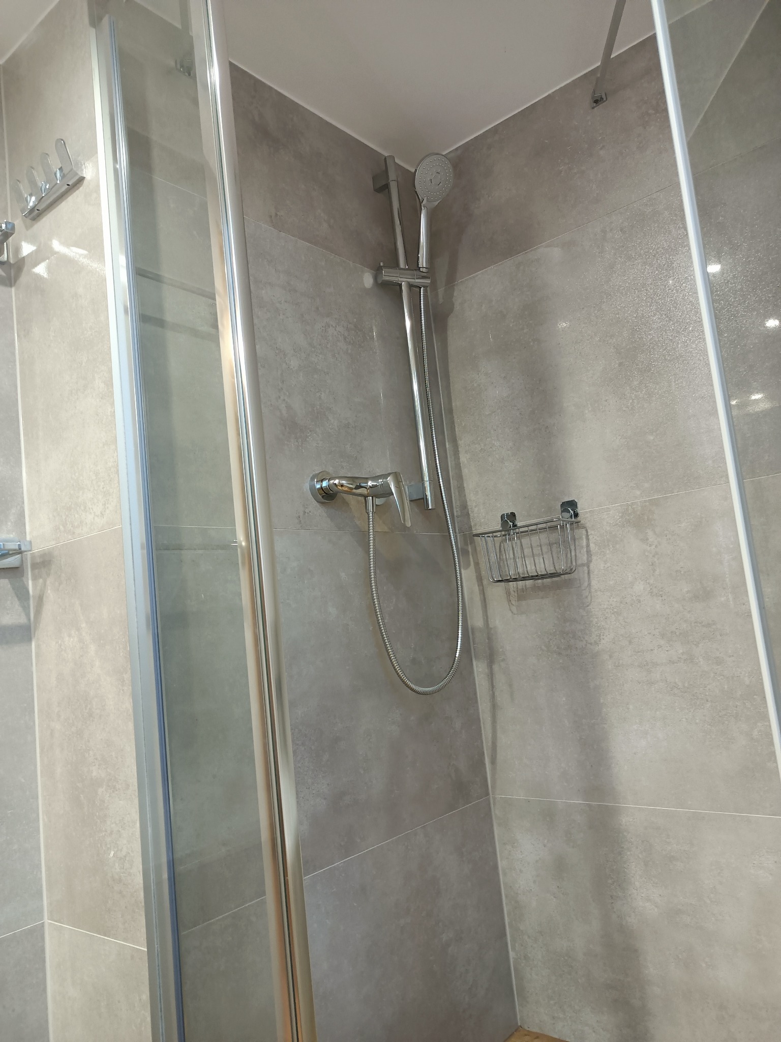 Kabina prysznicowa z szarymi płytkami imitującymi beton, chromowaną armaturą i półką na kosmetyki.