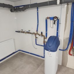 AquaXpert Sylwester Gajda - Biały zmiękczacz wody z niebieską górą, zamontowany w pomieszczeniu z szarymi rurami i grzejnikiem. Widoczne przyłącza i instalacja na białej ścianie z płytkami.