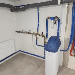 Biały zmiękczacz wody z niebieską g&oacute;rą, zamontowany w pomieszczeniu z szarymi rurami i grzejnikiem. Widoczne przyłącza i instalacja na białej ścianie z płytkami.