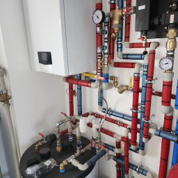 AquaXpert Sylwester Gajda - Biały piec Vaillant i rozbudowana instalacja hydrauliczna z rurami w izolacji czerwonej i niebieskiej, manometry, zawory. Widok z góry na zbiornik.