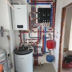 AquaXpert Sylwester Gajda - Kompletna instalacja grzewcza z kotłem Vaillant, zbiornikiem, rozdzielaczem i rurami w kolorach czerwonym i niebieskim. Widoczne manometry i zawory.