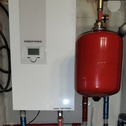 AquaXpert Sylwester Gajda - Biały piec grzewczy Defro Optima z czerwonym zbiornikiem wyrównawczym i rurami w izolacji, zamontowany na tle surowej ściany. Widoczne zawory i przyłącza.