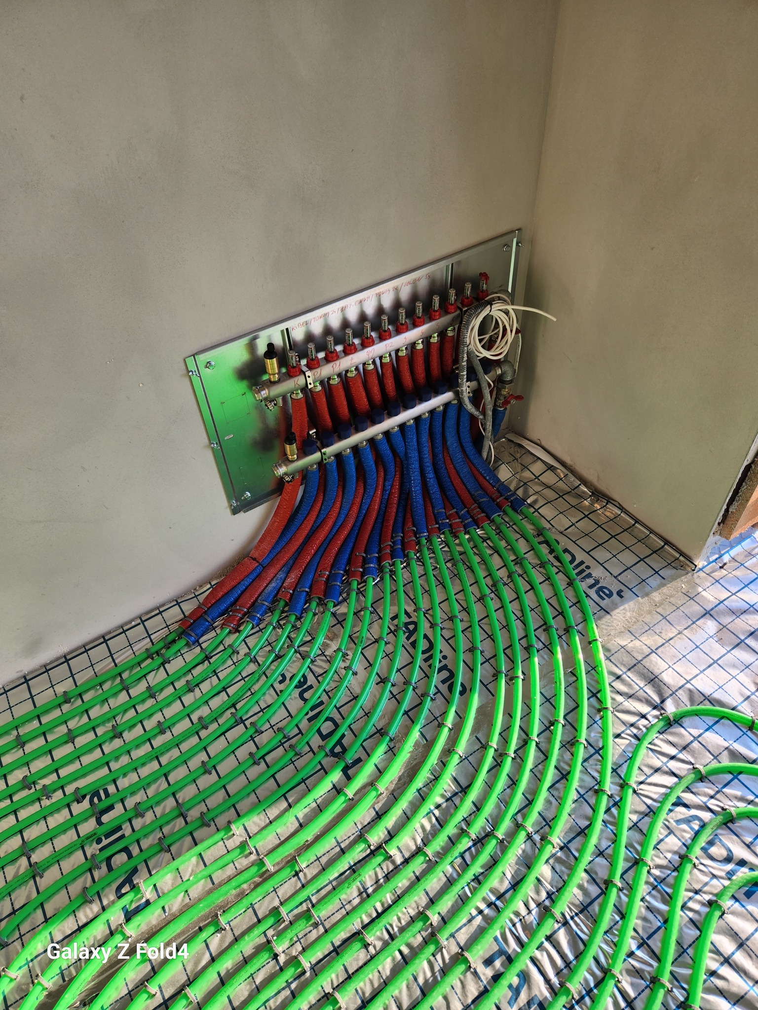 Rozdzielacz ogrzewania podłogowego z rurami w kolorach: czerwonym, niebieskim i zielonym, ułożonymi na folii z rastrem, zamontowany przy szarej ścianie.