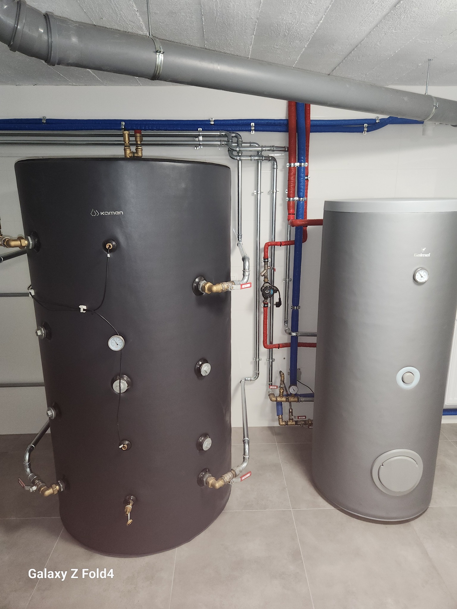 Dwa zbiorniki wody użytkowej (czarny i srebrny) z podłączonymi rurami miedzianymi i plastikowymi na tle białej ściany i szarej podłogi.