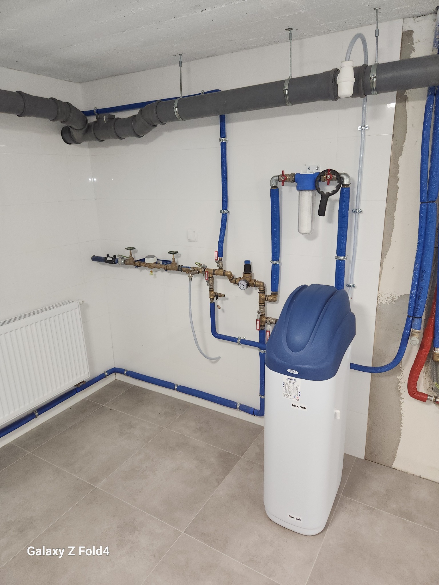 Biały zmiękczacz wody z niebieską górą, zamontowany w pomieszczeniu z szarymi rurami i grzejnikiem. Widoczne przyłącza i instalacja na białej ścianie z płytkami.