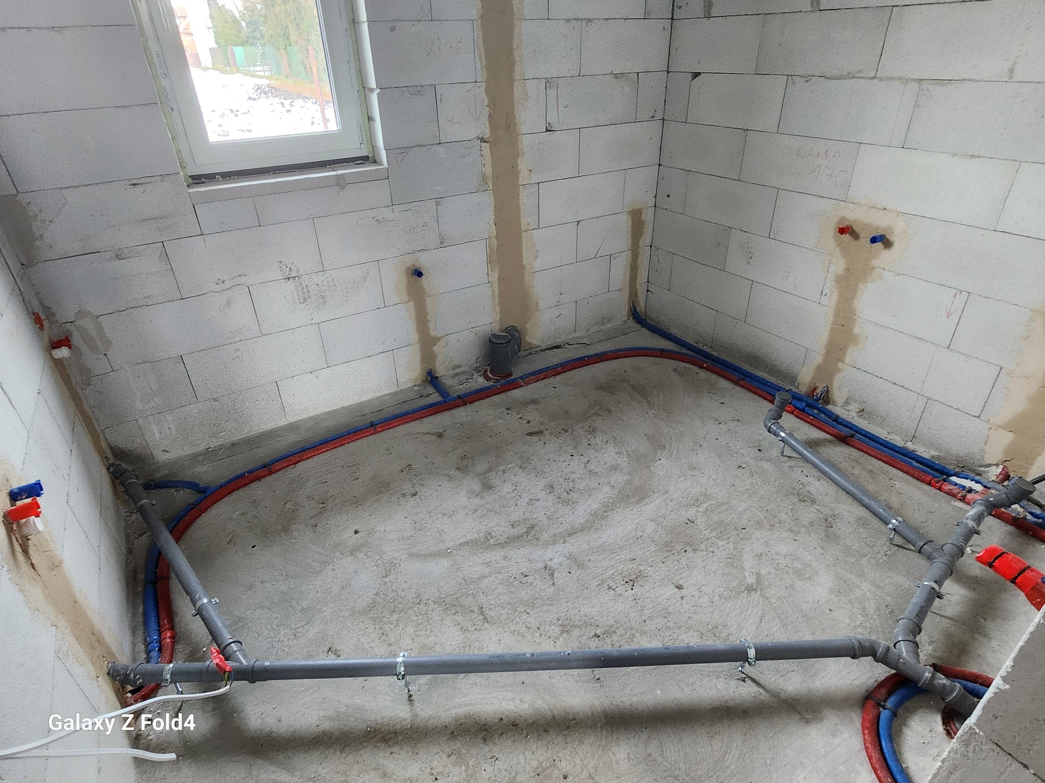 Surowy pokój z instalacją hydrauliczną: rury czerwone i niebieskie na podłodze, szare rury kanalizacyjne, ściany z białego betonu komórkowego z oknem.