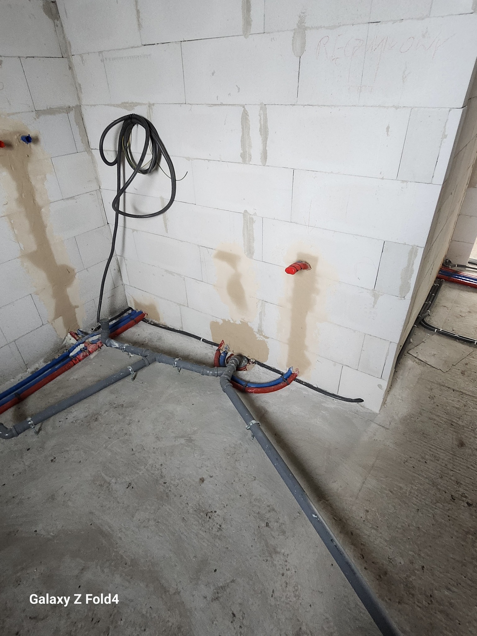 Surowe wnętrze z instalacją hydrauliczną: szare rury, czerwone i niebieskie złączki na tle białych ścian z betonu komórkowego. Widoczne przewody elektryczne.