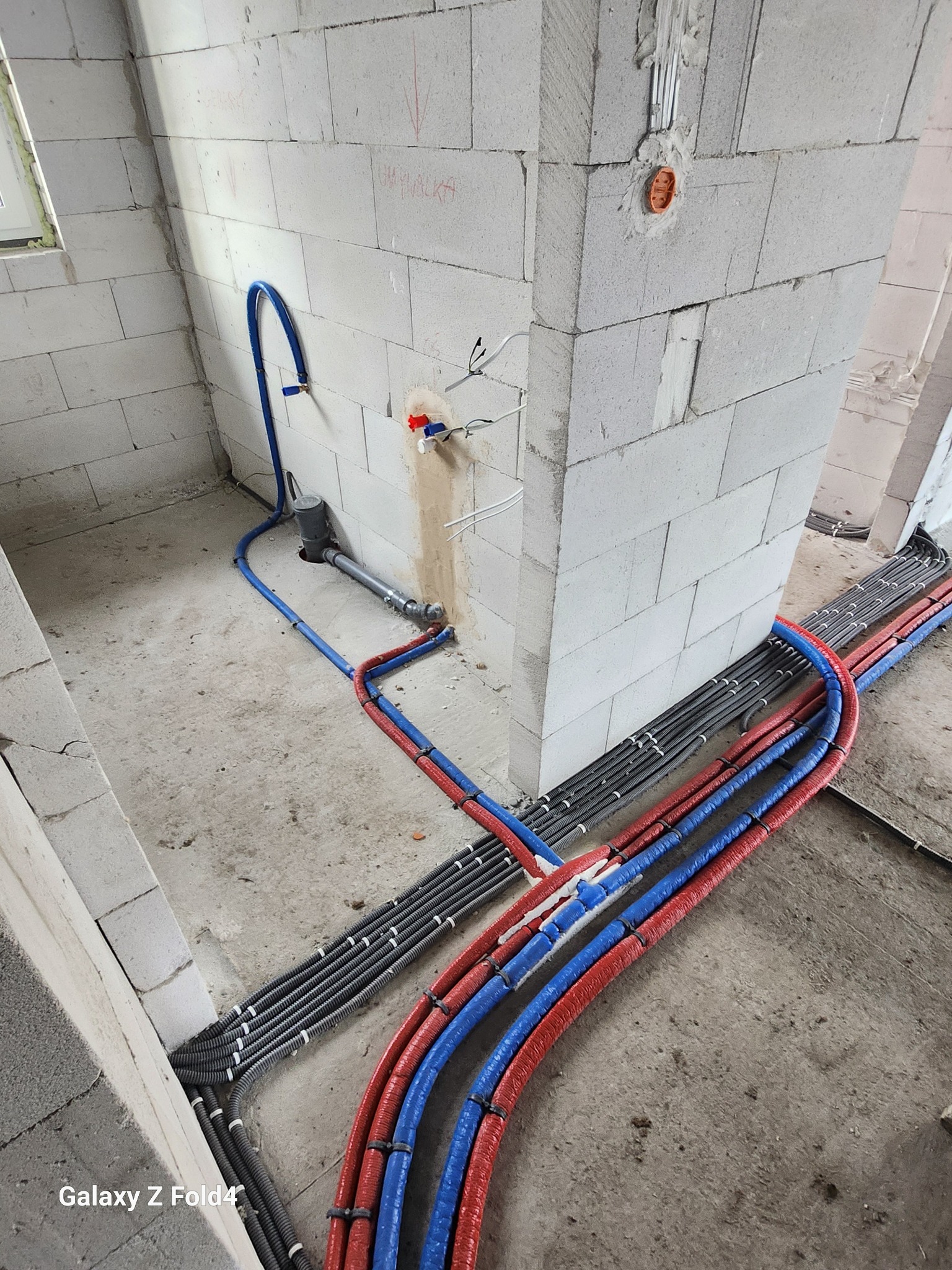 Instalacja rur w kolorach niebieskim i czerwonym, ułożonych równolegle na podłodze w surowym wnętrzu z bloków. Widoczne kable elektryczne i fragment okna.